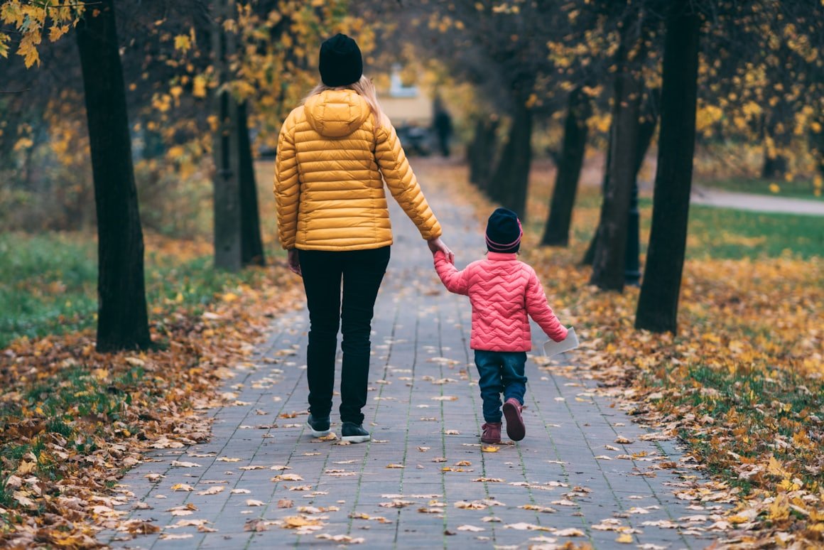Mutter und Kind gehen Hand in Hand eine Herbstallee entlang, beide von hinten